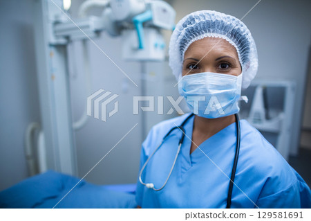 Female doctor standing in radiology suite wearing blue scrubs by X-ray machine with surgical mask 129581691