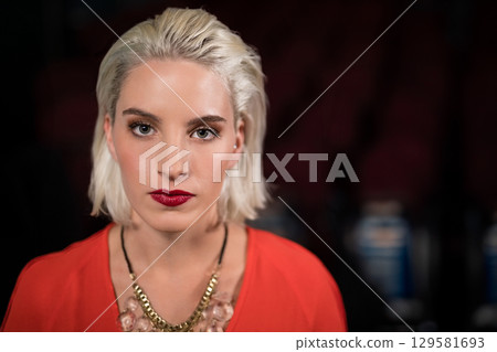 Woman gazing at camera posing in dark auditorium wearing red blouse and gold-and-pink necklace 129581693
