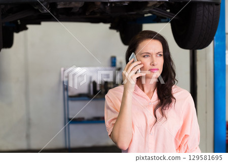 Woman calling on smartphone beneath raised car in repair bay and checking undercarriage, copy space 129581695