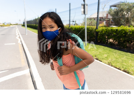 Girl student standing on sidewalk beside street with wire fence wearing teal backpack and blue mask Girl student standing on sidewalk beside street with wire fence wearing teal backpack and blue mask 129581696