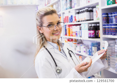 Female pharmacist standing in pharmacy aisle holding prescription slip near medicine shelves 129581700
