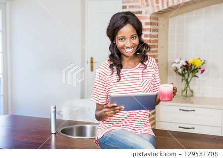 African American woman sitting on wooden countertop in kitchen holding tablet and pink coffee mug 129581708