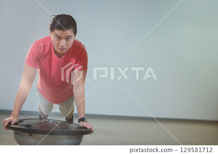 Adult Chinese woman performing push-up on half-dome trainer wearing sportswear in fitness studio 129581712