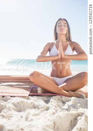 Woman wearing white swimsuit practicing yoga sitting cross-legged on bright mat on beach with waves Woman wearing white swimsuit practicing yoga sitting cross-legged on bright mat on beach with waves 129581716