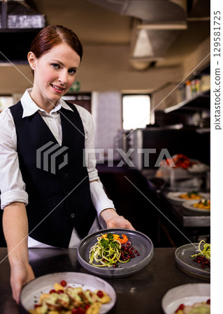 Female chef wearing uniform plating spiral veg salads with pomegranate seeds in kitchen, copy space 129581725