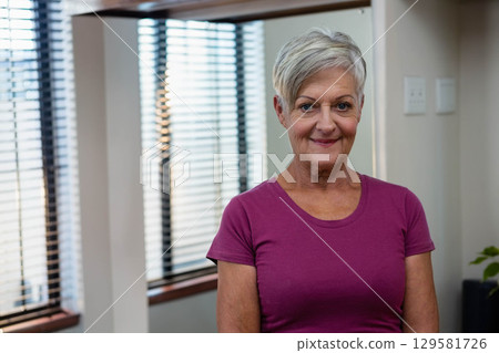 Senior woman standing facing camera in brightly lit room with wooden window blinds and potted plant 129581726