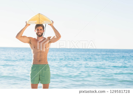 Man holding yellow surfboard overhead and standing waist-deep in ocean at sandy beach 129581728
