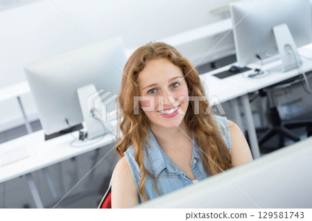 Female student smiling and using desktop computer at white desk in computer lab wearing denim shirt 129581743