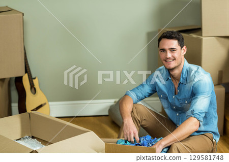 Man sitting on hardwood floor unpacking folded clothes from moving boxes near rolled rug and guitar Man sitting on hardwood floor unpacking folded clothes from moving boxes near rolled rug and guitar 129581748
