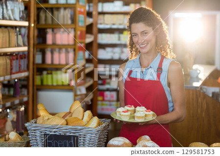 Woman in her twenties holding green plate of frosted cupcakes at bakery counter with bread loaves Woman in her twenties holding green plate of frosted cupcakes at bakery counter with bread loaves 129581753