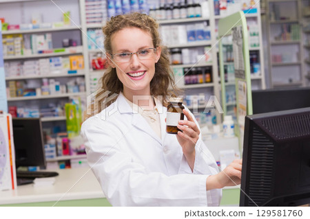 Female pharmacist in white coat holding brown medicine bottle behind pharmacy counter with monitor Female pharmacist in white coat holding brown medicine bottle behind pharmacy counter with monitor 129581760