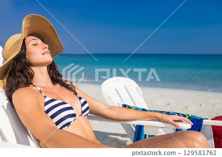 Woman in her twenties reclining on beach chair wearing bikini, straw hat, beach towel, copy space 129581762