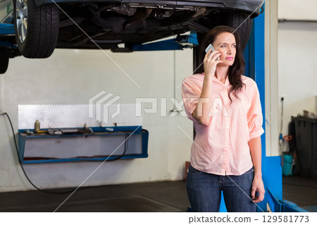 Woman standing in auto garage holding smartphone and examining raised car chassis on hydraulic lift Woman standing in auto garage holding smartphone and examining raised car chassis on hydraulic lift 129581773