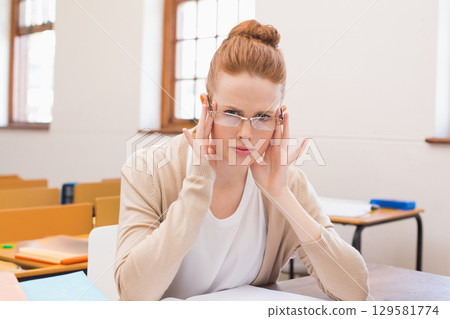 Female student adjusting eyeglasses while sitting at desk in classroom with open notebook, folders 129581774