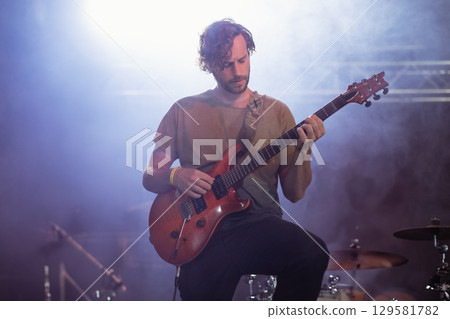 Male musician playing electric guitar on raised stage near drum kit under blue spotlights with fog Male musician playing electric guitar on raised stage near drum kit under blue spotlights with fog 129581782