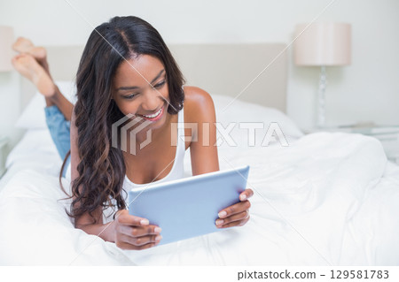 African American woman lying on bed in bedroom using tablet with white bedding and table lamp 129581783