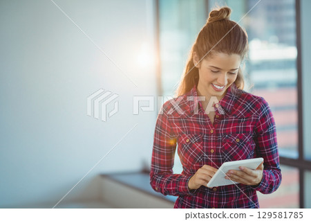 Woman in plaid shirt standing near large windows tapping tablet in office sun flare, copy space Woman in plaid shirt standing near large windows tapping tablet in office sun flare, copy space 129581785