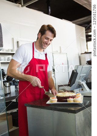 Male barista wearing red apron slicing chocolate cake behind cafe counter beside frosted cupcakes 129581786