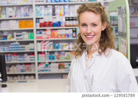 Smiling female pharmacist working behind counter, stocked shelves, pen in pocket, copy space 129581789