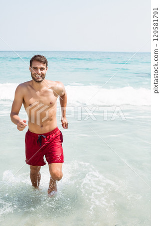 Man running from shallow ocean water onto sandy beach under clear sky wearing red swim trunks Man running from shallow ocean water onto sandy beach under clear sky wearing red swim trunks 129581791