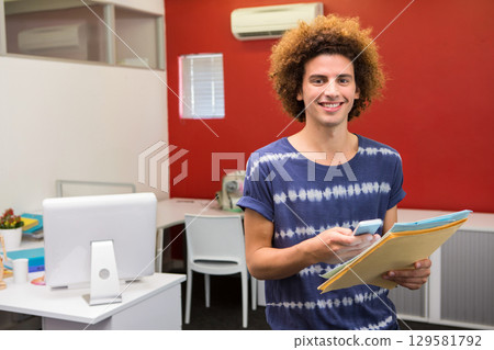 Curly-haired man standing in office holding smartphone and manila folders by monitor, copy space 129581792