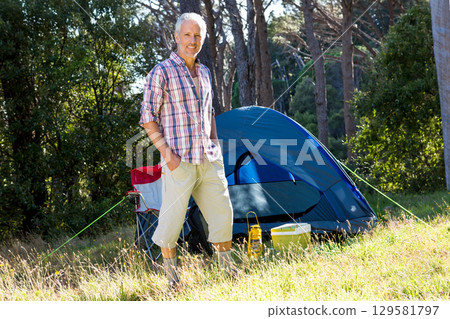 Senior man standing in sunlit clearing at forest edge beside blue tent with chair and cooler Senior man standing in sunlit clearing at forest edge beside blue tent with chair and cooler 129581797