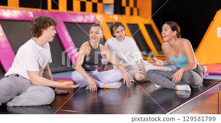 Group of enthusiastic young people sitting on trampoline and discussing upcoming jumping workout in entertainment center Group of enthusiastic young people sitting on trampoline and discussing upcoming jumping workout in entertainment center 129581799