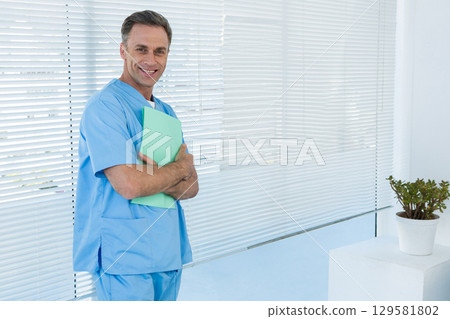 Middle-aged male wearing scrubs standing in office holding green folder beside plant, copy space Middle-aged male wearing scrubs standing in office holding green folder beside plant, copy space 129581802