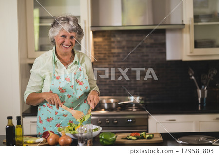 Senior woman tossing salad in white bowl with wooden spoon at kitchen island with sliced cucumbers Senior woman tossing salad in white bowl with wooden spoon at kitchen island with sliced cucumbers 129581803
