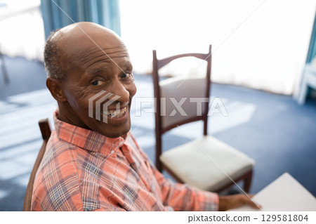 Senior African American man smiling while holding paper on chair in lounge wearing plaid shirt Senior African American man smiling while holding paper on chair in lounge wearing plaid shirt 129581804
