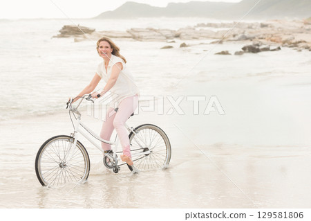 Woman riding white cruiser bicycle through shallow water along wet sandy beach with rocky outcrops Woman riding white cruiser bicycle through shallow water along wet sandy beach with rocky outcrops 129581806