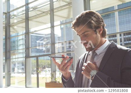 Man speaking into smartphone while wearing dark suit near glass walls in office lobby, copy space 129581808