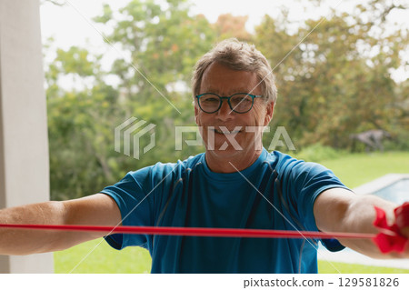 Senior man wearing teal shirt stretching red resistance band on patio overlooking lawn and pool Senior man wearing teal shirt stretching red resistance band on patio overlooking lawn and pool 129581826