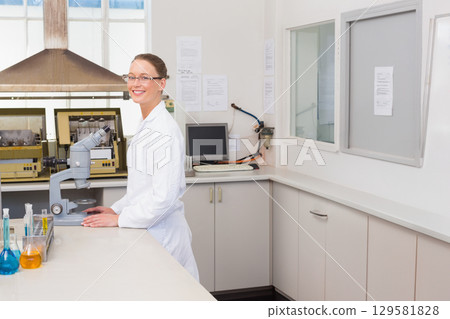 Female scientist wearing lab coat and glasses, working with microscope and test tubes in laboratory 129581828