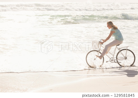 Woman riding white bicycle with front basket skimming surf along sandy beach barefoot, copy space 129581845