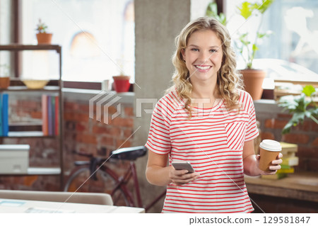 Teenage girl standing in loft-style workspace holding smartphone and paper coffee cup, copy space 129581847
