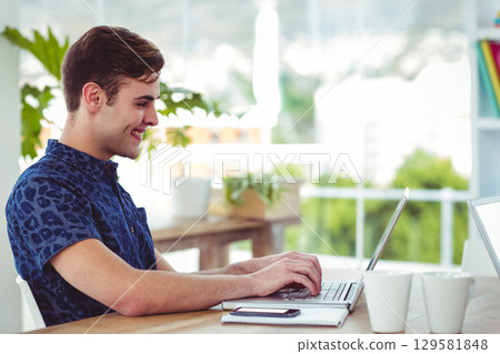 Man in early twenties wearing shirt typing on laptop at home office with smartphone, copy space 129581848