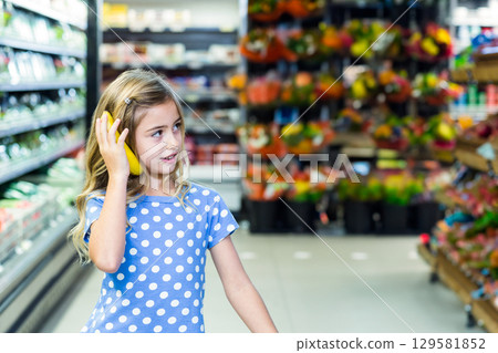 Female child holding banana like phone in supermarket produce aisle with flower displays 129581852
