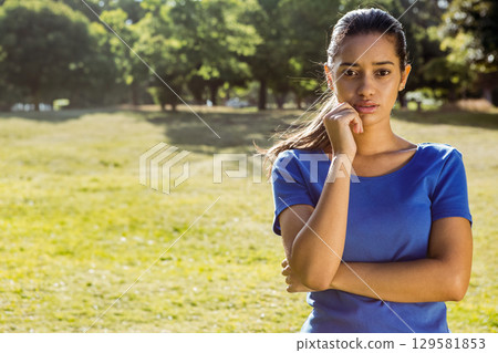 Woman wearing blue T-shirt standing in sunny park meadow thinking with hand on chin, copy space 129581853
