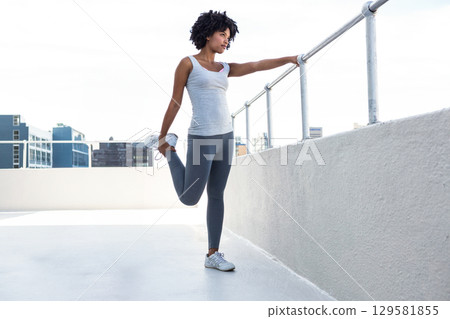 African American woman performing quad stretch on rooftop terrace using metal railing and sneakers 129581855