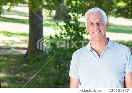 Senior man standing in sunny park under tree wearing grey polo shirt, copy space 129581857