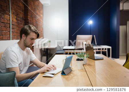 Man working on tablet and keyboard at wooden communal table in coworking space with coffee cup Man working on tablet and keyboard at wooden communal table in coworking space with coffee cup 129581876
