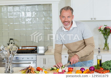 Middle-aged man chopping vegetables using chef knife on cutting board in home kitchen, copy space 129581879