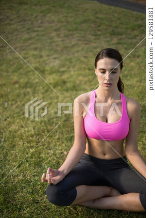 Fit woman meditating cross-legged on grass at park in pink top and dark leggings, copy space 129581881