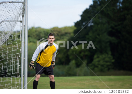 Male goalkeeper standing by goal with netting on pitch wearing yellow jersey, gloves, copy space 129581882