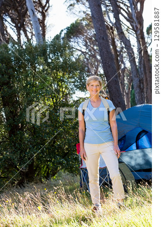 Senior woman standing in forest campsite wearing hiking backpack beside blue tent and camping chair 129581887