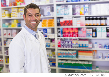 Male pharmacist smiling near meds shelves in pharmacy wearing lab coat and tie, copy space 129581888