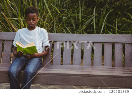 African American boy sitting on wooden bench in garden, reading bright green book, copy space 129581889