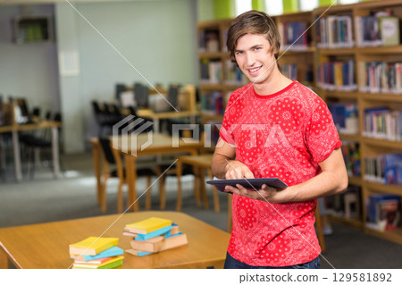 Man standing at wooden table in library reading room holding tablet near book stacks, copy space 129581892