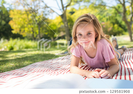 Child girl reading open book on red and white striped blanket at grassy park, copy space 129581908
